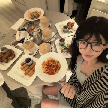 A young woman with glasses taking an overhead selfie at a table filled with various dishes including fries, spring rolls, salads, and coconut drinks.
