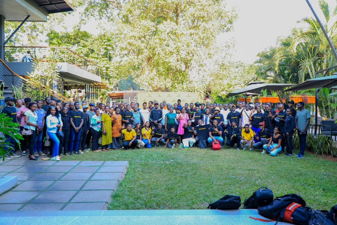 A large group of over 200 attendees poses for a group photo outdoors on a grassy lawn with tropical trees in the background, following the Binance Traders Masterclass meetup in Kampala focused on TradFi and Binance stocks.