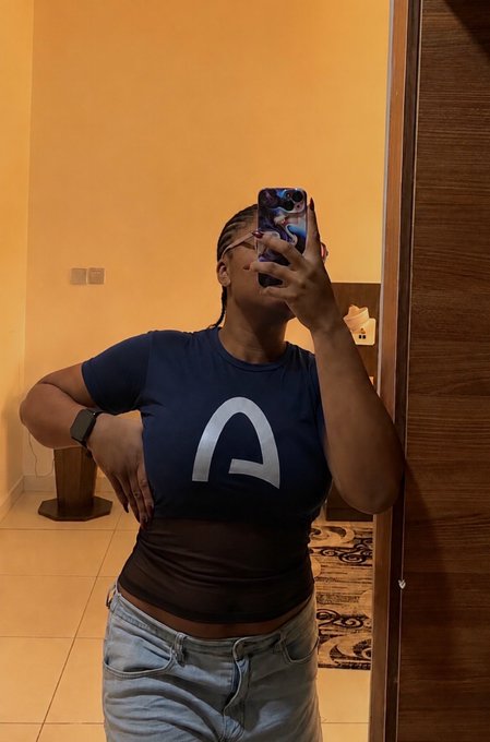 A woman takes a mirror selfie holding a phone with a blue flame design, wearing a navy blue t-shirt with a white "A" logo and light jeans. The "A" likely represents Arc, the blockchain network developed by Circle, tying into her post about Circle Developer Grants funding builders on Arc solving real-world payment challenges across Africa.