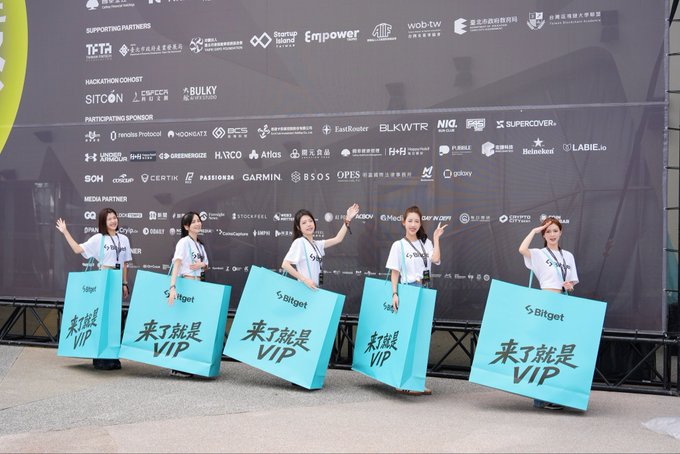 Five promotional models holding 'Bitget' bags with Chinese text '来了就是VIP' (Come and be VIP) at an event in front of a sponsor board.