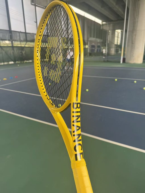 A yellow Yonex tennis racket stands upright on a green tennis court with tennis balls scattered in the background.
