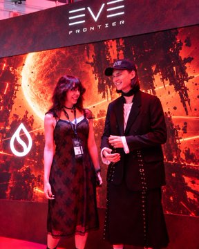 Two people pose in front of a 'EVE FRONTIER' banner featuring red and orange space-themed artwork, with one person in a black dress and the other wearing a black cap and long coat.