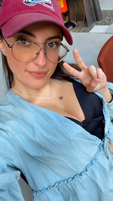 A selfie of a young woman wearing a maroon baseball cap, round glasses, and a light blue top, making a peace sign with her hand.