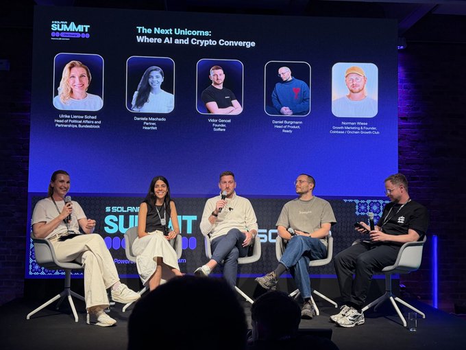 The image shows a group of six people sitting in chairs behind a stage, likely participating in a panel discussion or event related to AI and crypto. The audience appears engaged as they listen to the speakers' presentations. The backdrop features a large screen displaying images of various individuals, possibly representing different companies or organizations involved in the field of AI and cryptocurrency. The image also includes text that reads "The Next Unicorn: Where AI and Cryptocurrency Converge", indicating the main topic being discussed at this event.