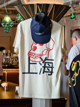 A cream-colored "上海" (Shanghai) T-shirt hangs on display featuring a red skull graphic, layered under a navy blue cap, in a stylish café setting.

The image ties to the "早安上海！" (Good morning Shanghai!) greeting by showcasing Shanghai-themed merchandise, likely from the Bored Ape community collaboration mentioned in the post.