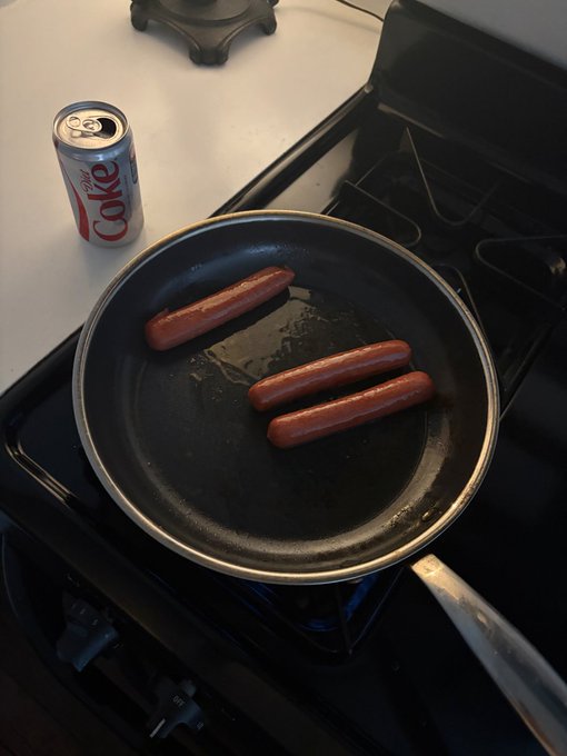 Four hot dogs cooking in a black frying pan on a gas stove, with a can of Diet Coke beside it on the counter.