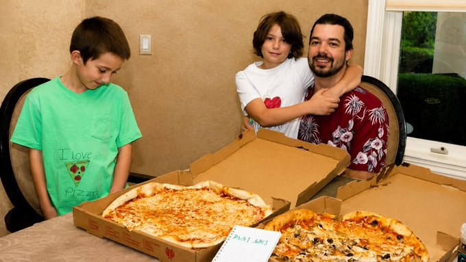 A family photo showing a father and two children gathered around a table with two large cheese pizzas in open delivery boxes, with one child wearing a green shirt that says 'I love pizza'.