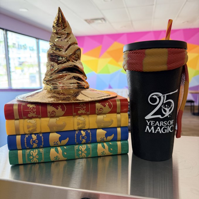A golden Sorting Hat rests atop a stack of four colorful Harry Potter-themed books beside a black tumbler featuring a striped Gryffindor scarf design and the text "25 YEARS OF MAGIC," celebrating a Harry Potter-themed café's silver anniversary with wizardly merchandise that any Muggle would love to spend their money on.