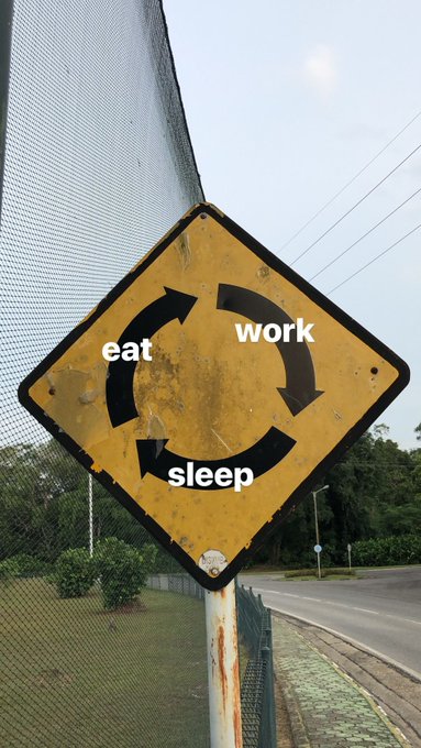 The image features a yellow sign with black text that reads "Eat, Work, Sleep". It appears to be located near a fence or gate, possibly at an intersection where people are crossing the street. The sign's message seems to encourage people to prioritize their health and well-being by taking breaks from work and getting enough sleep.
