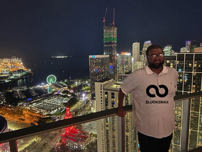 A man wearing a white "$LOOKSMAX" branded polo shirt poses on a high-rise balcony overlooking the illuminated Miami skyline at night, with the Port of Miami's glowing Ferris wheel and a red-lit historic building visible below. The elevated vantage point and nighttime cityscape reinforce the post's "we made it" sentiment of achieving success.