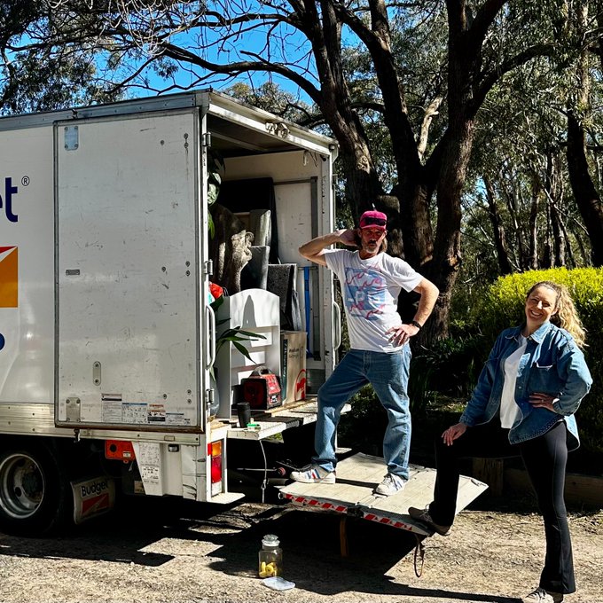 A man in a white t-shirt and pink cap stands on the lowered ramp of a Budget rental truck while a woman in a denim jacket poses beside him in a wooded outdoor setting.