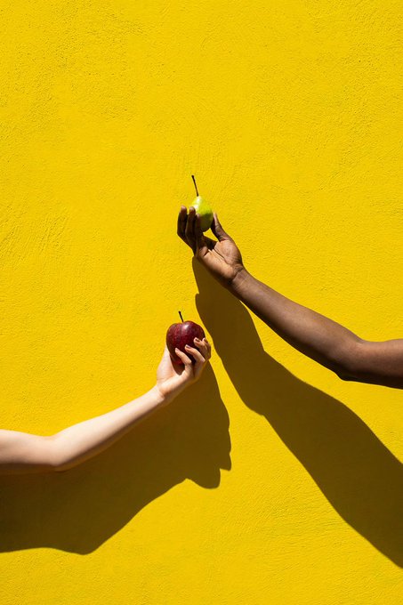 Two hands, one light-skinned and one dark-skinned, exchanging fruits against a bright yellow wall—one hand holding a green pear and the other holding a red apple, with strong shadows cast on the wall.