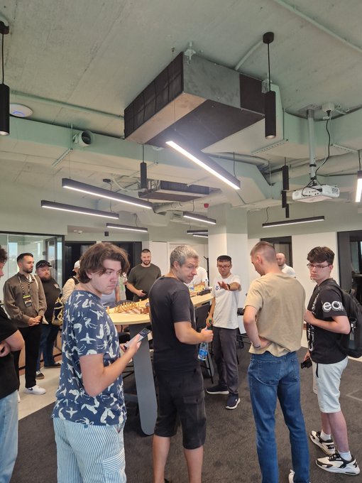A group of people gathered in a modern office space with exposed ceiling pipes and linear pendant lights, including a person wearing a shirt with 'eai' visible on the backpack.