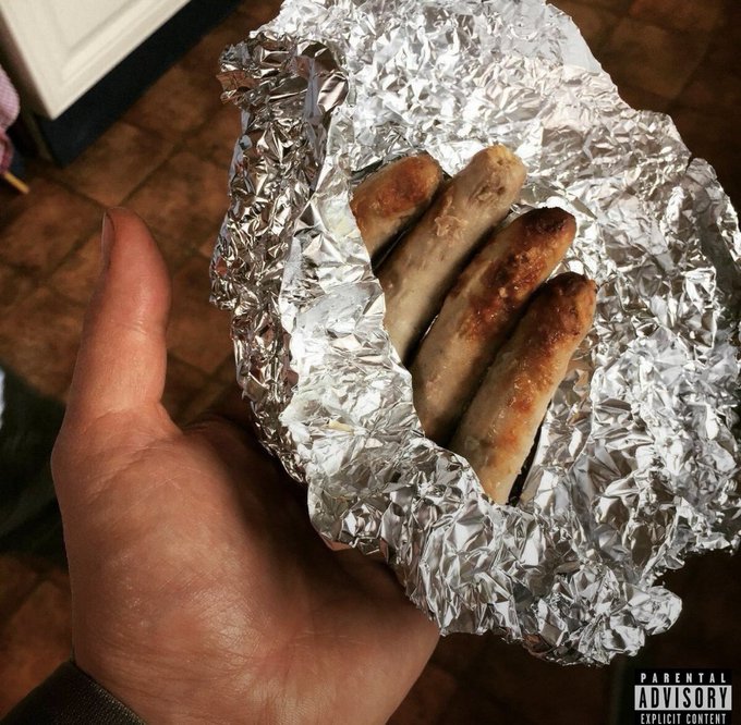 A hand holding aluminum foil cradling what appears to be three cooked sausages, with a 'Parental Advisory Explicit Content' label overlaid.