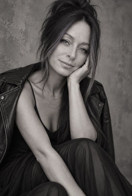 Black and white portrait of a woman with long dark hair wearing a leather jacket, resting her chin on her hand against a textured gray background.