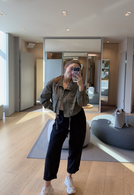 A woman takes a mirror selfie in a modern, well-lit room, wearing a khaki jacket, black pants, white sneakers, and sunglasses.