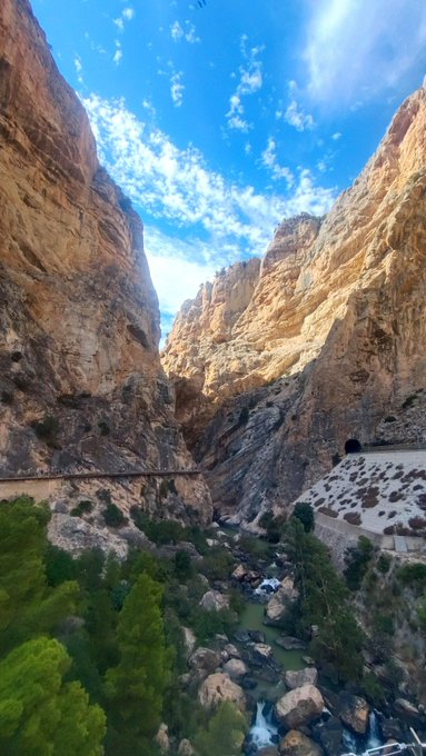 A scenic view looking down into a deep canyon with rocky walls, a small stream, and green vegetation, with a road visible on the left and a tunnel entrance on the right.