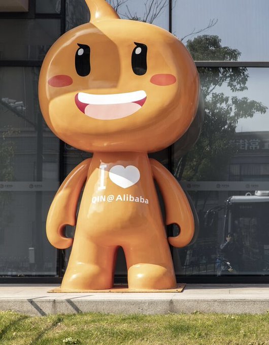 An orange Alibaba mascot statue with the text 'I ❤️ QIN @ Alibaba' stands on a sidewalk in front of a glass building.