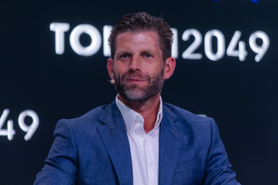 A clean-shaven man in a blue checkered blazer and white shirt speaks into a lapel microphone at the TOKEN2049 conference, with the event's logo visible behind him. The image appears to show Eric Trump during his appearance alongside Zach Witkoff and Zak Folkman of World Liberty Financial on the OKX Main Stage at TOKEN2049 Singapore.