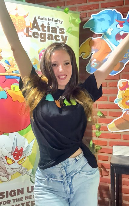 A woman poses enthusiastically with arms raised in front of a promotional banner for "Axie Infinity: Atia's Legacy," which features colorful Axie character artwork. The setting appears to be at a branded event related to the post's mention of building within the Ronin Network and the Web3 gaming community.