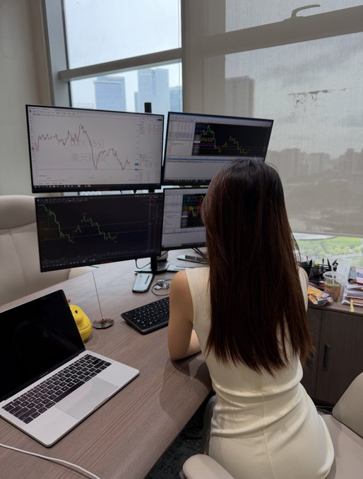 A woman with long dark hair sits at a desk with four monitors displaying stock trading charts, alongside a laptop, in a high-rise office with city views.