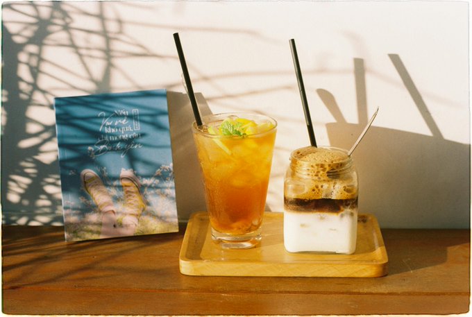 The image features a wooden table with two drinks placed on it - one of them being a tall glass filled with tea, while the other has ice cream in it. The table also displays a book titled "Good morning everyone New week. New sleep goals. New rewards." and another book that says "@quipnetwork is still running their Quantum Echoes NFT campaign on NucleusCodes". These books are placed next to each other, suggesting they might be related or part of the same collection. The scene appears to be set in a room with a white wall as the backdrop for these items.