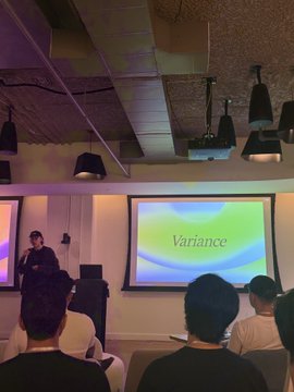 A presenter speaks to an audience in a dimly lit venue with a projector screen displaying the word "Variance," likely illustrating the statistical concept tied to the upside potential of @joinCero's reward-centric neobank benefits.