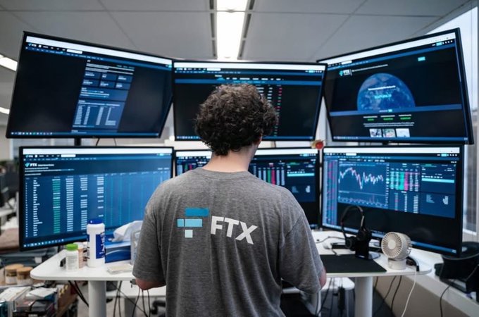 A person wearing a gray 'FTX' branded t-shirt sits at a workstation surrounded by six monitors displaying financial charts, data tables, and trading interfaces.