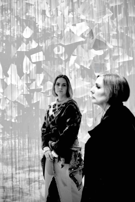 A black and white photograph showing two women standing in front of an artistic installation with hanging translucent papers, one woman in a leopard print cardigan looking at the camera while another in a dark coat stands in profile.