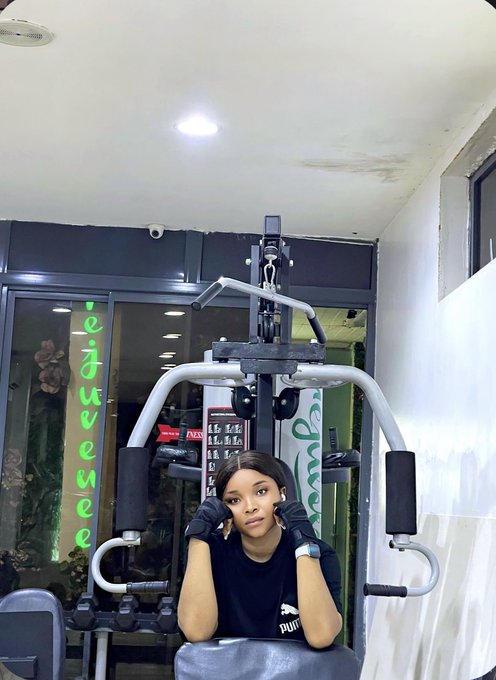 A woman wearing a black t-shirt and black gloves poses on a gym machine, with a 'Juvenne' sign visible in green neon lighting in the background.