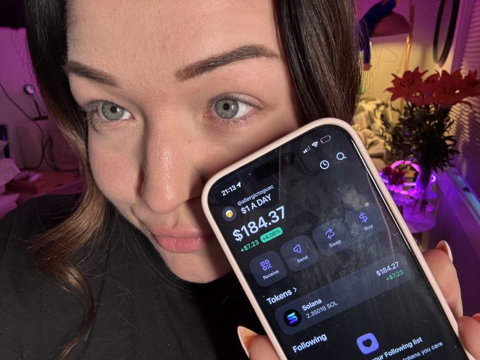 A young woman holds up her phone displaying a Solana wallet with a balance of $184.37 and 2.3516 SOL, visually documenting day 224 of her "$1 a day until ATH" SOL accumulation challenge.
