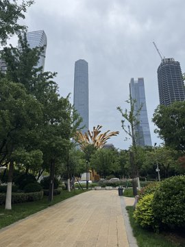 The image shows a city street with tall buildings, including skyscrapers, lining both sides of the road. The sky above appears cloudy, creating an urban atmosphere. In the foreground, there are trees that provide some greenery amidst the concrete structures. A sidewalk runs along one side of the street, and a statue is located on the other side. The post accompanying this image provides information about the location or event taking place in the city.