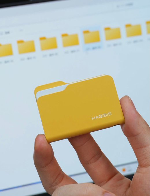 A hand holds a small yellow card holder branded 'HAGIBIS' in front of a computer screen displaying yellow folders.