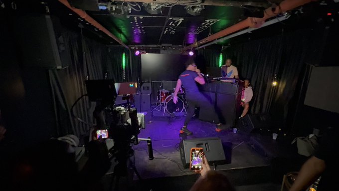 A performer sings on a dimly lit venue stage with purple and green lighting while an audience member records the show on their smartphone.