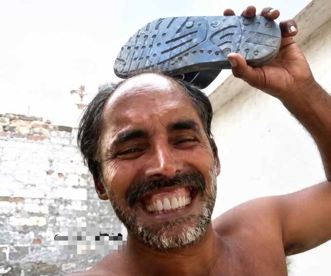 A shirtless man with a mustache and beard smiles broadly while holding up a sandal over his head, with a weathered brick wall in the background.