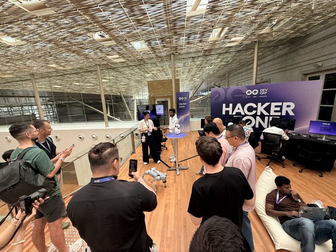 A group of people gathered in a modern indoor venue with wooden floors and a perforated metal ceiling, watching a robot demonstration near a purple banner reading 'HACKER ZONE'.