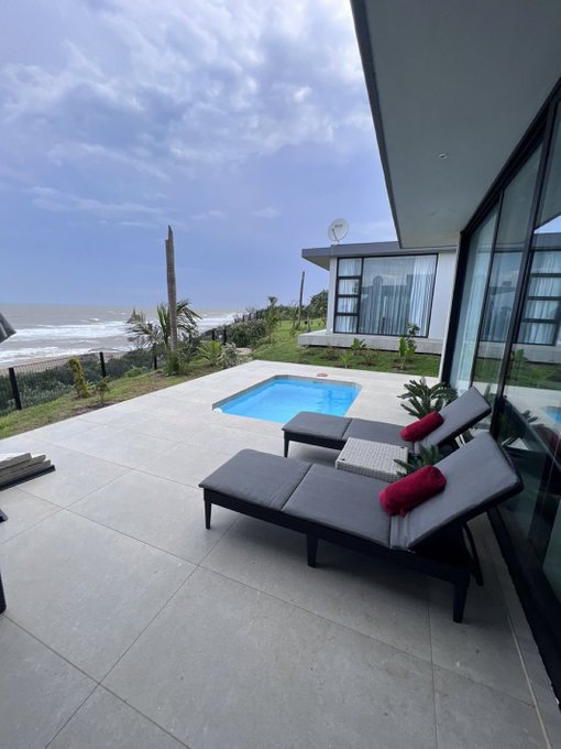 An oceanfront patio featuring a small swimming pool, two lounge chairs with rolled red towels, and a view of the beach and cloudy sky.