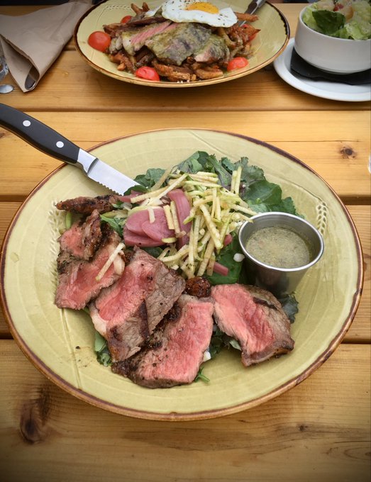 A green plate holds sliced rare steak over greens, topped with thin fried potatoes, pickled red onions, and accompanied by a small metal cup of dressing, with a second plate of food visible in the background on a wooden table.