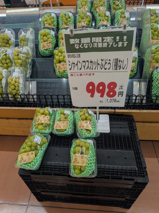 Shiny Muscat grapes (シャインマスカットぶどう) are displayed in plastic clamshell containers at a Japanese market, with a sign announcing "数量限定!! なくなり次第終了します" (Limited quantity! Available until sold out) and a price of 998 yen (1,078 yen with tax) per bunch.

The caption "I agonized over it, but... I couldn't buy it" relates to the image perfectly—these premium Japanese Shine Muscat grapes are notoriously expensive, and at 998 yen per bunch, the steep price likely made the shopper pause, weighing whether the luxurious treat was worth the splurge before ultimately deciding to pass.