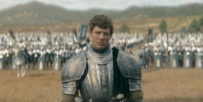 A man in ornate armor stands in a field with an army in the background, appearing to be a scene from a medieval fantasy film or TV series.
