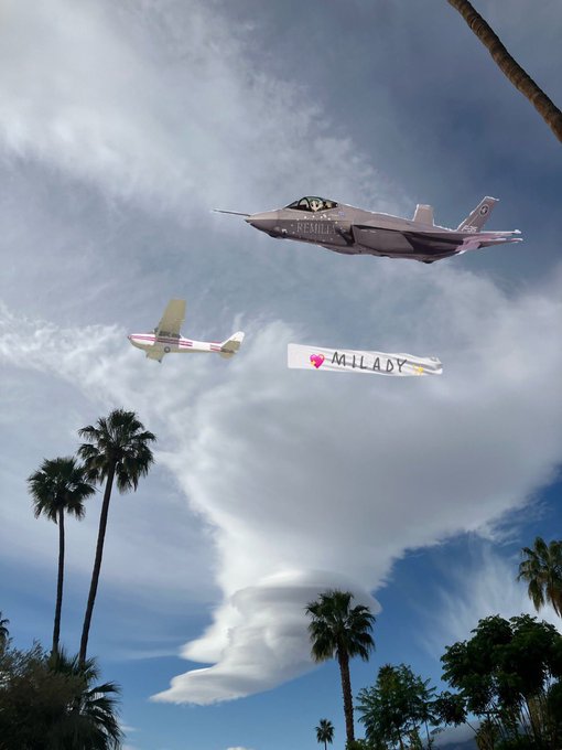 A photo of a fighter jet and a small plane flying in formation while pulling a banner that reads 'MILADY' with a heart and sun icon, set against a cloudy sky with palm trees visible below.