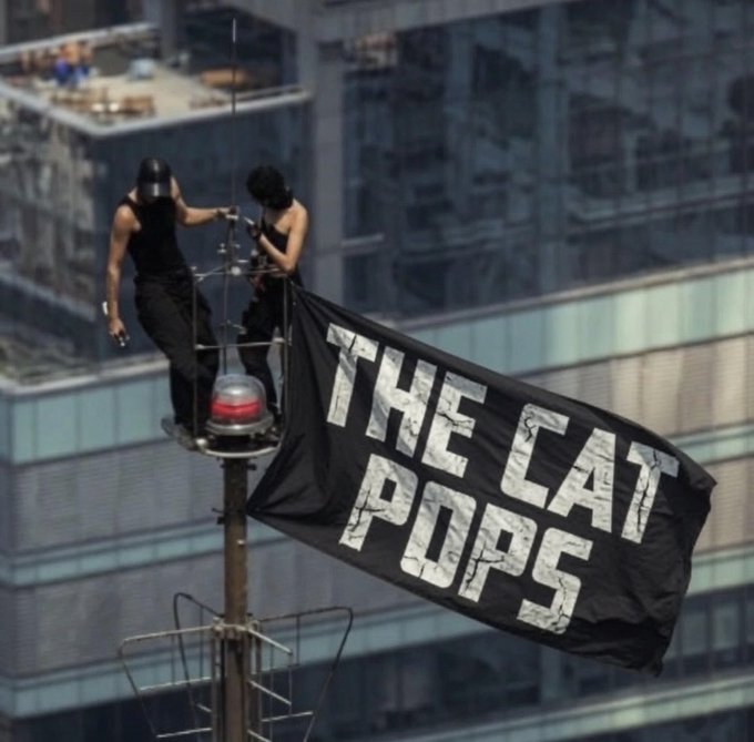 Two people in black clothing stand on a high antenna platform, attaching a black flag that reads 'THE CAT POPS' in white distressed lettering.