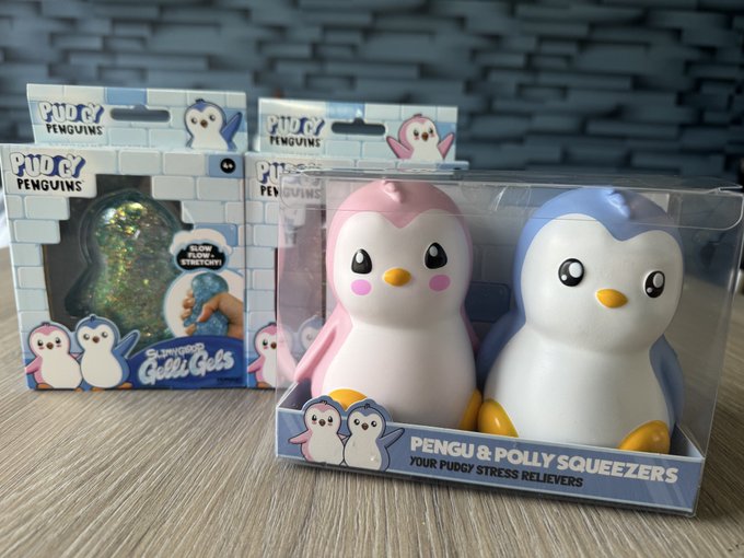 Boxed Pudgy Penguins stress reliever squishy toys sit on a surface, with one package prominently labeled "PENGU & POLLY SQUEEZERS YOUR PUDGY STRESS RELIEVERS" and another showing a "GLOW FLOW STRESS BALL" and "SQUISHY QUIET GELS".