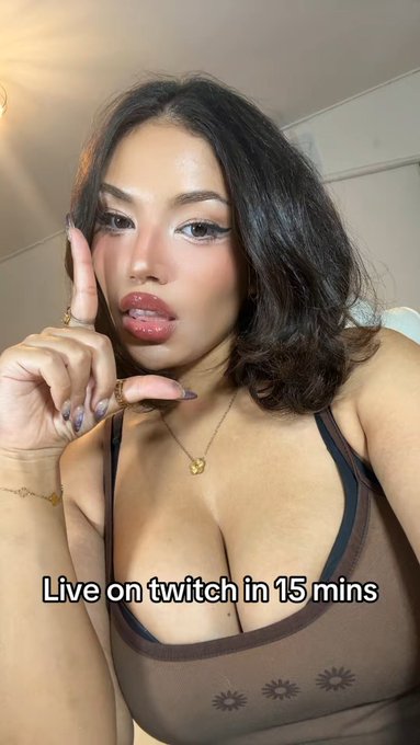 A selfie of a woman with dark hair, heavy makeup, and long manicured nails wearing a brown tank top with sun designs, pointing upward; overlaid text reads "Live on twitch in 15 mins".