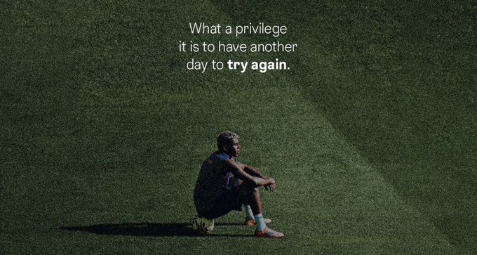 A soccer player sits alone on a vast green field in shadow, with the text "What a privilege it is to have another day to **try again**." overlaid in white. The image's message of perseverance and new beginnings aligns with the post's theme of starting the day fresh, hinting at an opportunity (likely a giveaway) for the community to "try again" at their luck or fortunes.