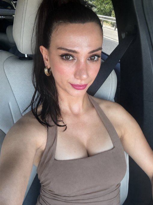 A woman with dark hair pulled back, wearing a brown halter top and gold hoop earrings, takes a selfie in the driver's seat of a car. The image captures a confident, glowing look that complements the cheerful "Gm frens!" morning greeting, radiating the bright, energetic vibe of Hump Day.