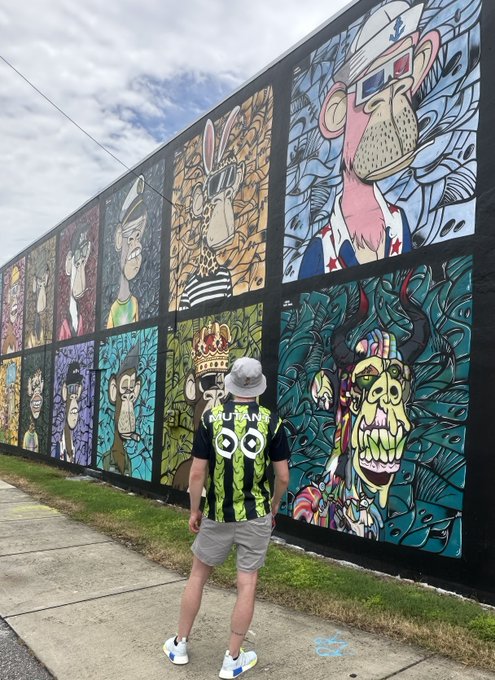 A person in a watermelon-patterned shirt and bucket hat stands before a vibrant mural featuring a grid of colorful cartoon monkeys styled like Bored Ape Yacht Club NFTs, including one with a crown and another skull-faced "mutant" ape—visually capturing the "Mutant Apes are the alpha" caption by highlighting the graffiti's tribute to the mutant variant of the iconic NFT collection.