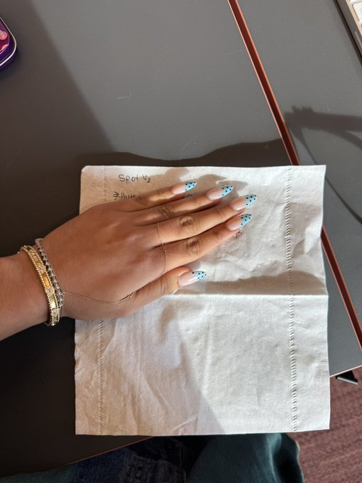 A hand adorned with a gold bracelet and blue polka dot nail art rests on a crumpled napkin with "Spot V2" and "Julian" written on it, matching the post about a "napkin session for @jup_spot V2" while showcasing the updated manicure.
