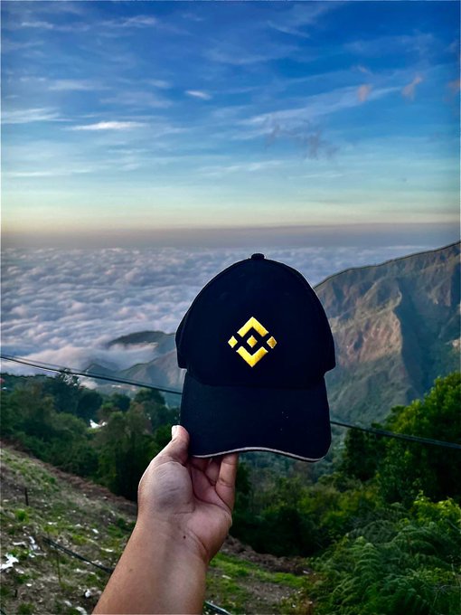 A hand holds up a black baseball cap featuring a yellow Binance logo against a scenic mountain backdrop with clouds filling the valley below.