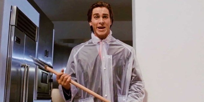 Actor Christian Bale, appearing as Patrick Bateman from American Psycho, stands in a modern kitchen wearing a clear plastic garment cover and holding an axe.